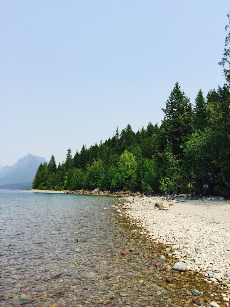 Tranquil view of Lake McDonald's rocky shoreline with lush forests and distant mountains.